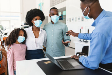 Obraz premium Car purchase during covid lockdown. Afro family in face masks taking electronic car key from salesman at auto dealership. Black clients buying or renting new vehicle at showroom shop