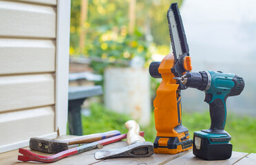 A collection of power tools on a wooden surface. Includes a cordless drill, a saw, a wrench, and a level. Background features greenery and a blurred fence.