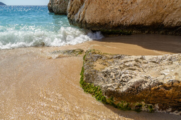 : Scenic view of turquoise sea wave crashing on a golden sandy beach with coastal rocks. Clear water and bright sunlight on the Mediterranean shore. September, Antalya region, Turkey.