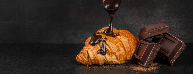 Chocolate sauce pouring over croissant with dark chocolate pieces on black background