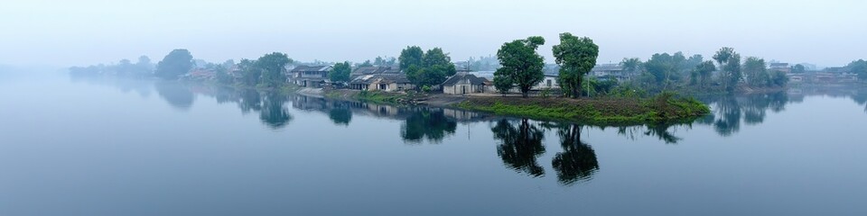 Obraz premium Serene riverside village at dawn with misty reflections and lush greenery