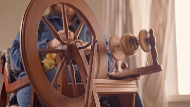 Spinning wheel being used by a person to turn raw fleece and fiber into strands of yarn.  Wooden traditional home tool used to transform raw materials into supplies for making clothing.