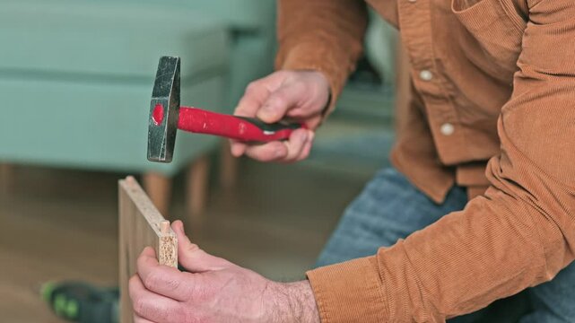Skilled individual using a hammer to drive a dowel into a wooden panel, demonstrating precise woodworking techniques in a well-lit indoor environment with visible furniture
