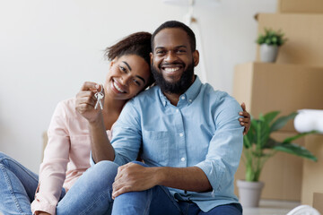 Fototapeta premium Glad cheerful young african american family show key and enjoy purchase of real estate in room with cardboard boxes with things and plants, copy space. Celebrating moving in new house, relocation
