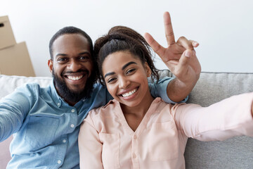 Smiling young black couple rest on couch, man shows peace sign, makes selfie in room with cardboard...