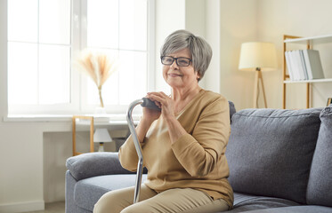 Happy senior woman with cane sitting alone on living room sofa at home and smiling. Confident...
