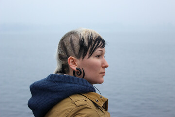 Natural, unretouched portrait of a young woman with an original shaved hairstyle and earring, standing by the sea. Authentic lifestyle image representing individuality, real beauty.  © Светлана Мищенко