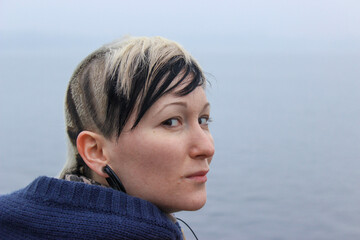 Natural, unretouched portrait of a young woman with an original shaved hairstyle and earring, standing by the sea. Authentic lifestyle image representing individuality, real beauty.  © Светлана Мищенко