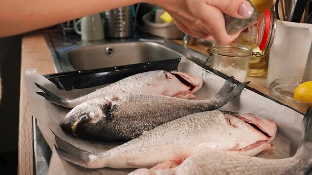 Woman season whole sea bream with salt and pepper on baking tray, prepare fresh, healthy homemade seafood meal for dinner or family gathering.Real life kitchen routine and everyday lifestyle moment.