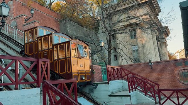 Historic Budav&aacute;ri Sikl&oacute; funicular railway car ascending Castle Hill in Budapest, Hungary, on a sunny autumn day, passing by another carriage with beautiful classical architecture in the background.

