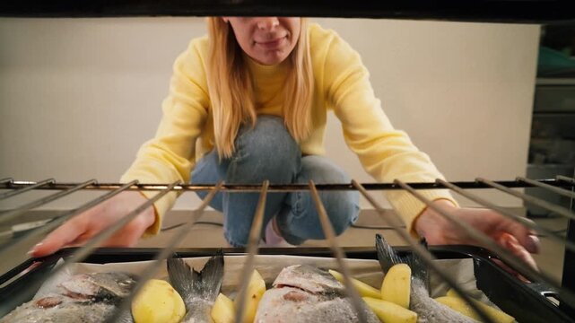 Young woman slides tray of fresh dorado and sliced potatoes into preheated oven, pov from inside appliance showing home cooking and healthy meal preparation.Healthy homemade food, everyday cooking