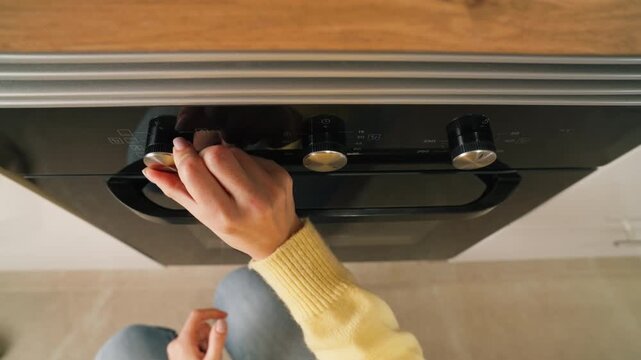 High angle view of woman open oven door and set temperature and cooke mode with control knobs on front panel before baking food in  modern kitchen at home.