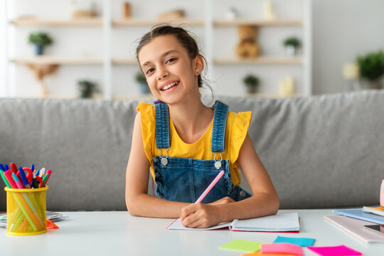 Modern Education Concept. Portrait of smiling little female student sitting at desk in living room, writing notes in her notebook or diary, looking posing at camera, selective focus
