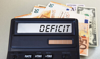 DEFICIT text written on a calculator next to the background of euro banknotes. Finance or business concept.