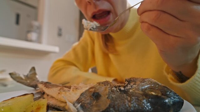Woman in mustard sweater delicately pulling flaky meat from baked gilthead bream with fork, savoring fresh, delicious seafood meal at table.Cozy lifestyle and homemade meal concept.