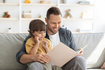 Naklejka premium Story time. Loving father reading interesting fairy tales book to his little son, interested adorable boy enjoying time with his daddy, sitting together on sofa at home