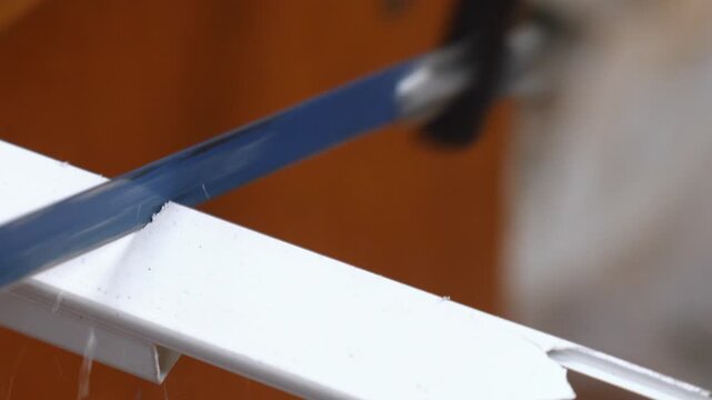 Close-up view of a hacksaw blade precisely cutting through a white plastic cable trunking, with small plastic particles flying off during the sawing process in a workshop environment