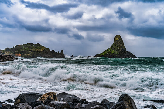 Dramatic coastal scene featuring rough waves crashing against dark volcanic rocks in Sicily. Two prominent sea stacks rise from the turbulent waters&mdash;one lush with greenery and a small orange building,
