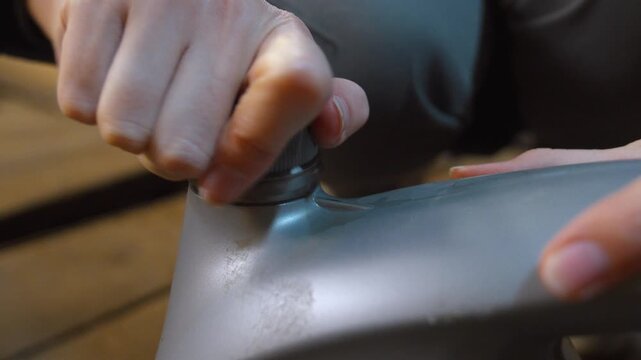Close-up of a woman's hands unscrewing the cap of a gray plastic canister, breaking the seal to open the container of engine oil before performing vehicle maintenance in a garage
