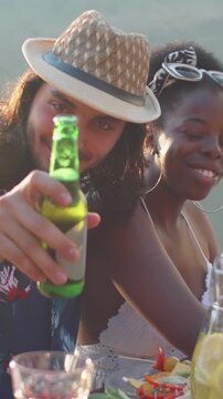 Vertical portriat shot of Middle Eastern man and his African-American girlfriend sharing joyful moment at table during lakeside gathering with friends, smiling for camera
