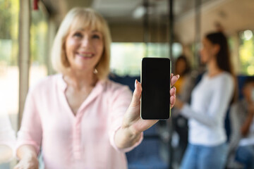 Great Offer. Excited mature lady taking bus holding cell phone with black blank screen in hand, showing device close to camera. Gadget with empty free space for mock up. Online ticket, gps navigation © Prostock-studio