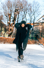 Fototapeta premium Smiling woman in a cozy black coat and fur hat walks joyfully through a snowy park, surrounded by trees and winter scenery, capturing the essence of winter fun and happiness