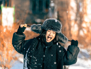 Young woman with dark hair wearing a furry hat and winter coat joyfully playing in the snow, surrounded by trees and a vibrant winter landscape, capturing the essence of winter fun
