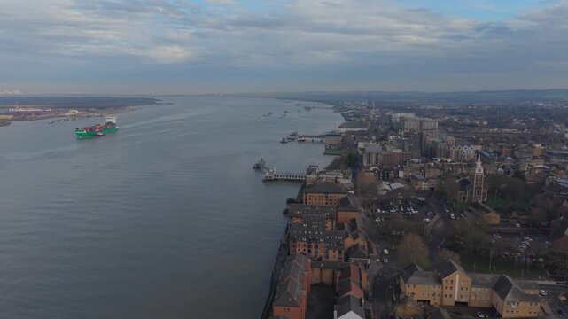 Container ship on the river Thames sailing by Gravesend town. Aerial view of large green vessel escorted by pilot boat towards the Docks near London. Gravesend town with historic church and pier.