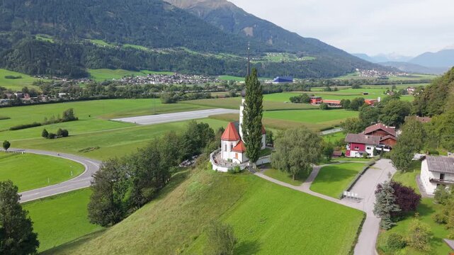 A small church with a pointed bell tower in a valley in Austria. Aerial view of the religious site against the backdrop of the mountains and landscape of the Oberes Inntal valley in Tyrol