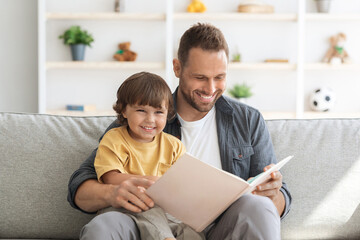 Naklejka premium Storytime. Young loving father reading fairy tales to his cute little son, sitting together with big child book on sofa at home and smiling