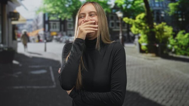 Young woman covering mouth with hand on sunlit cobblestone street, wearing black turtleneck and crossed arms; shy amusement.
