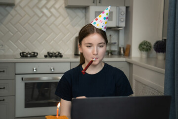 Young girl wearing a festive party hat and blowing a party horn, celebrating her birthday through a video call in the kitchen, surrounded by a birthday cake and candles