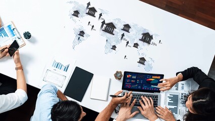A diverse business team collaborates over a laptop displaying financial data and market insights, surrounded by charts and a world map, illustrating strategic planning. Raster