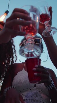 Vertical low angle shot of diverse group of men and women clinking their glasses in cheerful toast, dancing at their summer lake party, gathered on pier at dusk