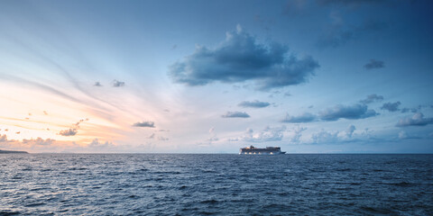 Fototapeta premium Open sea at dusk with a passenger ship sailing on the horizon under dramatic clouds, shown in a wide panoramic composition with ample copy space for travel and maritime concepts.