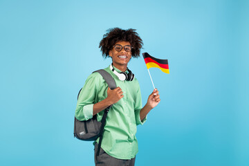 Modern education and knowledge exchange. African american teen guy in glasses with backpack, headphones and small Germany flag, posing on blue background, copy space