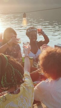Vertical high angle shot of diverse and joyful group of friends, gathered at dinner table on wooden pier for lake party, raising their glasses in toast