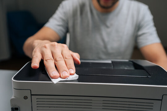 Close-up cropped shot of man performing printer maintenance, wiping dust off office laser printer, ensuring technology hygiene, cleaning and servicing electronic equipment in home office.