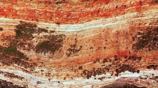 Rock formations show distinct layers of sediment and minerals on a cliff. The colors range from brown to red with white streaks. Plants grow in some areas, highlighting the natural environment.