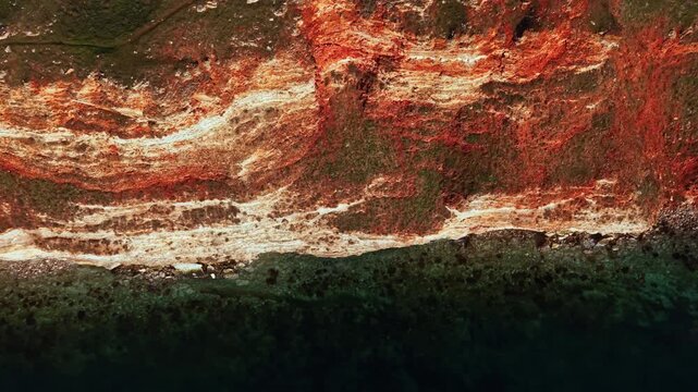 Cliffs rise above the water, displaying layers of color and texture. Waves gently lap at the base, and rock formations create a dramatic outline against the sky.