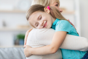 Naklejka premium Family Love Concept. Cute small girl hugging tight her smiling grandmother, sitting on the couch in living room at home, expressing love and tenderness. Selective focus, blurred background