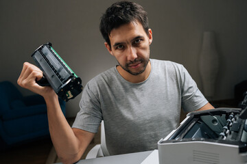 Young man holding empty toner cartridge from laser printer, looking directly at camera with frustrated expression while attempting to fix office equipment.