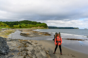 A hiker stands on a quiet beach in Russia, surrounded by a stunning landscape of cliffs and grassy...