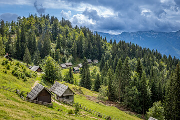 Planina Zajamniki shows wooden huts surrounded by trees on a hillside in Slovenia. Clouds gather over distant mountains creating a natural scene.