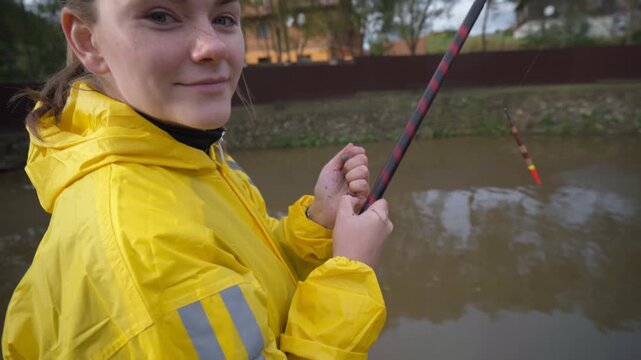 White woman yellow raincoat casting rod closeup, hands gripping rod, line threaded through guides, bobber visible on murky water, concentrated wrist movement,
