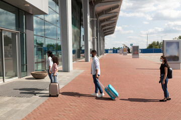 Travels During Pandemic. Side view of multiracial diverse passengers in medical masks walking with bags at airport building, standing in queue line at entrance. Social distancing due to covid outbreak