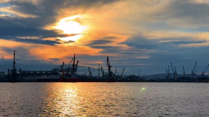 Sunset over the harbor with cranes and water reflections in the background. The sun sets over the harbor, casting colors across the water and silhouettes of cranes in the distance.