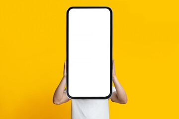Unrecognizable guy in white t-shirt hiding behind huge modern smartphone with white empty screen, posing on yellow studio background, mockup, copy space. Gadgets addiction concept