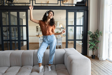 Young Caucasian woman in wireless headphones listening to music and dancing on sofa to favorite...