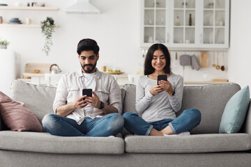 Device addiction, great mobile game, mobile app and social networks at spare time. Smiling young arab woman and man with smartphones typing messages and chatting with friends in living room interior